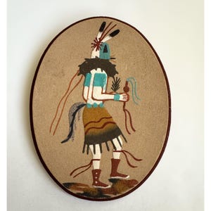 May include: An oval sand painting depicting a Native American figure. The figure is adorned with a headdress, turquoise and white clothing, and a brown skirt. The artwork features brown, blue, and tan colors, with a dark brown border.