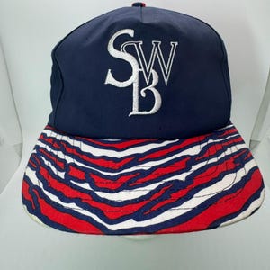 May include: A navy blue baseball cap with a white embroidered "SWB" logo. The brim features a red, white, and blue striped pattern. The cap has a classic, structured design.