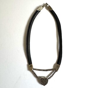 May include: Black leather choker necklace with silver-toned metal accents. The necklace features a heart-shaped pendant and decorative metal ends. The necklace is designed to be worn around the neck.
