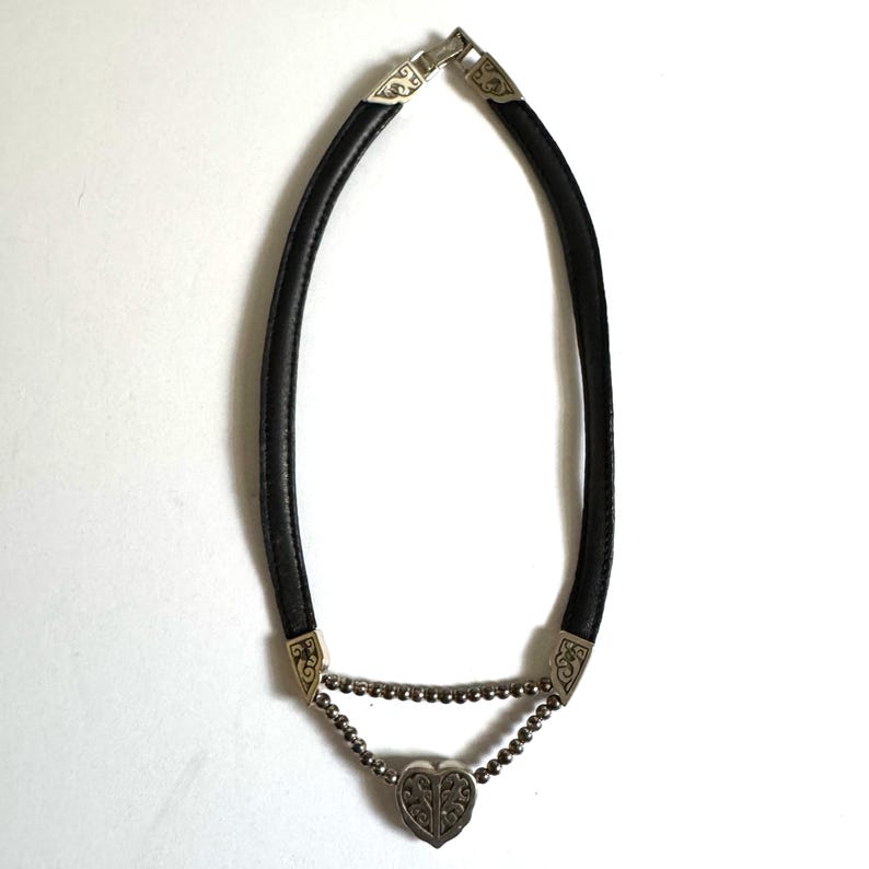 May include: Black leather choker necklace with silver-toned metal accents. The necklace features a heart-shaped pendant and decorative metal ends. The necklace is designed to be worn around the neck.