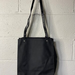 May include: A black tote bag with a black leather strap. The bag has a zipper closure and a flat base.