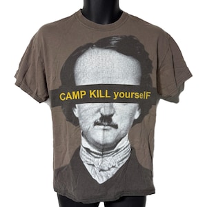 May include: A brown t-shirt featuring a black and white portrait of a man with a mustache. A yellow banner across the eyes reads "CAMP KILL yourself". The shirt has short sleeves and a crew neck.