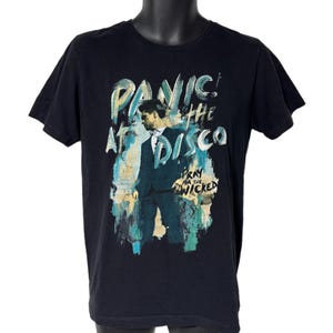 Könnte beinhalten: Schwarzes T-Shirt mit dem Text "Panic! At The Disco" in einer stilisierten Schriftart und einem Porträt einer Person im Anzug. Der Satz "Pray for the Wicked" ist ebenfalls auf dem T-Shirt zu sehen.