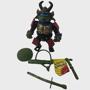 May include: A Teenage Mutant Ninja Turtle action figure in a samurai outfit, with accessories including a shield, sword, and banner. The banner reads "Sewer Samurai" with a sun graphic. The figure is green, blue, red, black, and gold.
