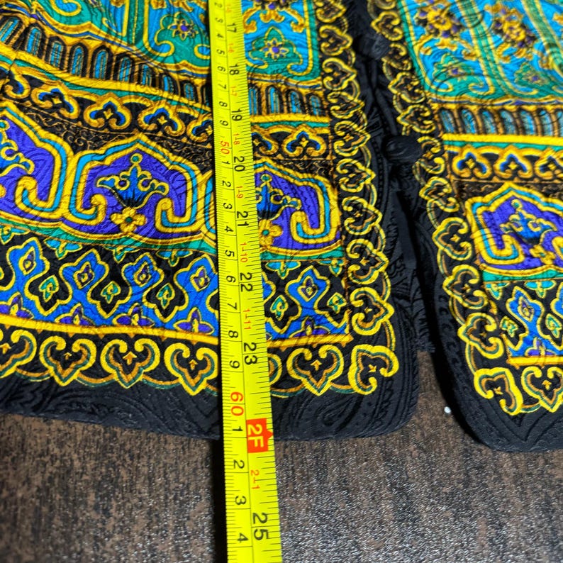 Pode incluir: Close-up de uma pe&ccedil;a de roupa colorida e estampada com acabamento preto. O design apresenta tons de verde-azulado, amarelo e roxo com um padr&atilde;o ornamental, tipo paisley. A pe&ccedil;a de roupa &eacute; medida com uma fita m&eacute;trica amarela.