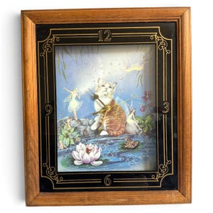 May include: A rectangular wall clock with a wooden frame and a black border featuring gold numerals 3, 6, 9, and 12. The clock face displays an illustration of a tabby cat sitting by a pond with fairies, a frog, and a water lily.