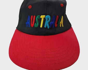 May include: Black baseball cap with a red brim. The word "AUSTRALIA" is embroidered across the front in colorful letters. The cap has a red button on top and red eyelets.