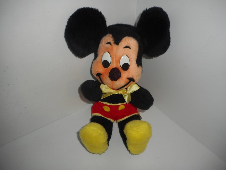 May include: A plush Mickey Mouse doll with black fur, large round ears, and a peach-colored face. He wears red shorts with yellow buttons, yellow shoes, and a yellow bow tie. The doll is sitting.