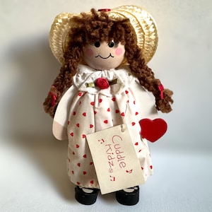 May include: A handcrafted doll with brown yarn hair, wearing a white dress with red hearts and a straw hat. The doll holds a red heart and a tag that reads "Cuddle Kidz". The doll has a painted face with rosy cheeks.
