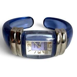 May include: A cuff watch featuring a translucent blue resin band and a rectangular face with a light purple dial. The watch includes silver-tone metal accents on both sides of the case, with the text Roberto Fiori and Quartz visible.
