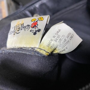 May include: A black nylon bag with a Mickey Mouse logo and the text "Disney Store" on the tag. The tag also says "Made in China, RN# 84167, 100% Nylon, Clean with Damp Cloth Only, Disney".