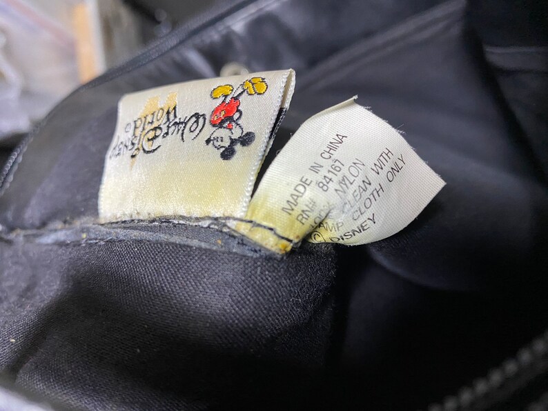 May include: A black nylon bag with a Mickey Mouse logo and the text "Disney Store" on the tag. The tag also says "Made in China, RN# 84167, 100% Nylon, Clean with Damp Cloth Only, Disney".
