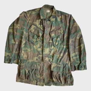 May include: A camouflage jacket with multiple pockets and a button-down front. The jacket has a collar and long sleeves. The camouflage pattern features shades of green, brown, and tan.