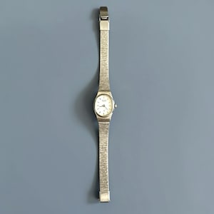May include: A silver-toned wristwatch with a white face and a mesh band. The watch has a small, oval-shaped face with black hands and markers. The band is made of a fine, woven metal. The watch is displayed against a solid blue background.