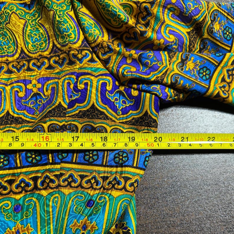 Pode incluir: Close-up de um tecido estampado com um design complexo. O tecido apresenta elementos amarelos, azuis, verdes e pretos. Uma fita m&eacute;trica amarela &eacute; colocada sobre o tecido, mostrando medidas em cent&iacute;metros.