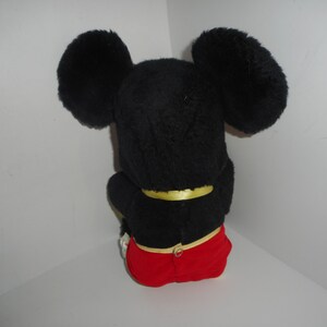 May include: A plush Mickey Mouse toy, viewed from the back. The toy features large, black, fluffy ears, a black body, and red shorts. A yellow band is around the waist. The toy is sitting down.