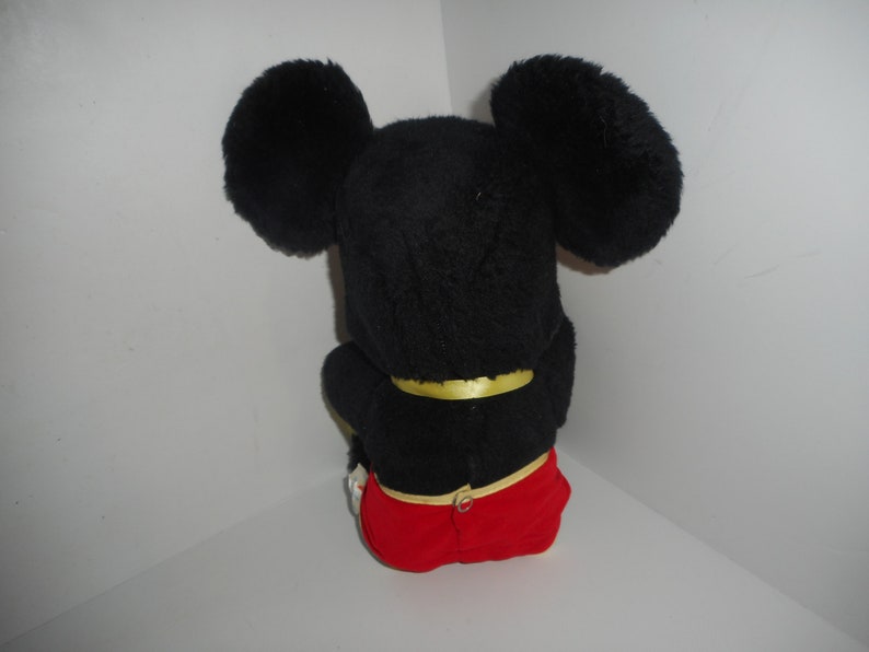 May include: A plush Mickey Mouse toy, viewed from the back. The toy features large, black, fluffy ears, a black body, and red shorts. A yellow band is around the waist. The toy is sitting down.