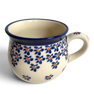 May include: A round ceramic mug with a cream-colored base featuring a repeating pattern of small blue flowers with orange centers. The mug has a thick, dark blue rim and a matching handle decorated with a single blue flower.