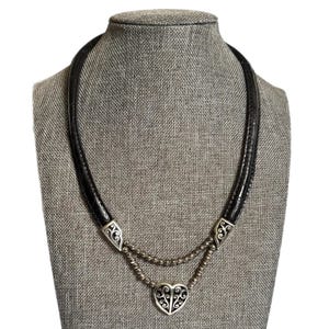May include: A black leather necklace with a silver heart pendant. The necklace features a double strand of small silver beads and decorative silver accents. The heart pendant has a filigree design, displayed on a grey mannequin.