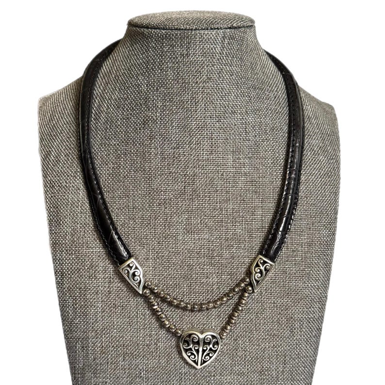 May include: A black leather necklace with a silver heart pendant. The necklace has a double strand of small silver beads and decorative silver accents. The heart pendant has a filigree design and is displayed on a gray mannequin.