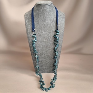 May include: A long necklace with teal-colored stone beads and a dark blue fabric cord. The beads are irregularly shaped and strung together. The necklace is displayed on a gray mannequin.