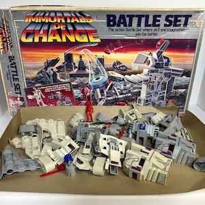 May include: Vintage "Immortals of Change" Battle Set toy box with various plastic pieces and figures. The box features the text "Immortals of Change" and "Battle Set". The set includes gray and off-white building components, a red figure, and other accessories.