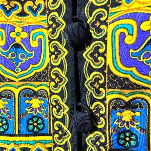 Pode incluir: Close-up de um tecido estampado com fundo preto. O design apresenta formas ornamentadas repetidas em amarelo, azul e verde-azulado. O tecido tem um fecho de bot&atilde;o no lado esquerdo.