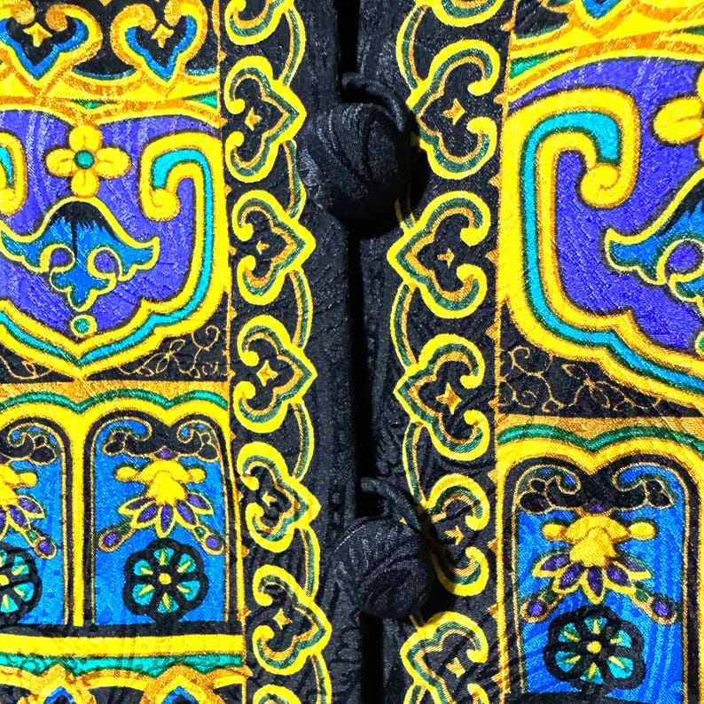 Pode incluir: Close-up de um tecido estampado com fundo preto. O design apresenta formas ornamentadas repetidas em amarelo, azul e verde-azulado. O tecido tem um fecho de bot&atilde;o no lado esquerdo.