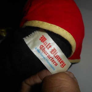May include: A plush toy with a red hat trimmed with gold and a black body. A white tag with red text reads "Walt Disney Characters" and "&copy; Walt Disney Productions".