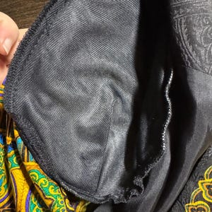 Pode incluir: Close-up do forro interior de uma pe&ccedil;a de roupa preta. O forro &eacute; um tecido liso e escuro, possivelmente cetim ou material semelhante. O exterior da pe&ccedil;a apresenta um padr&atilde;o paisley em ouro, amarelo e azul.