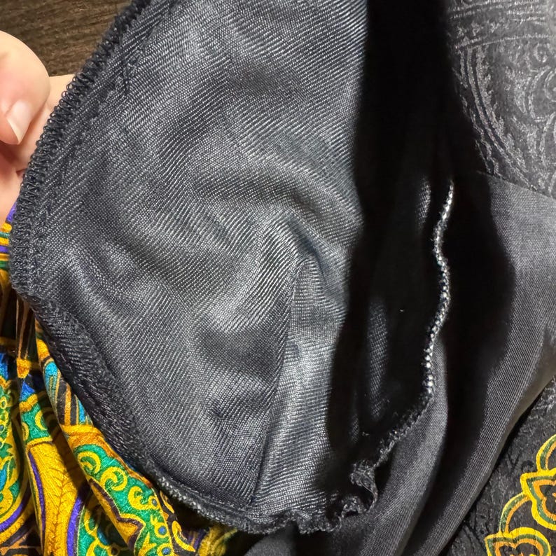 Pode incluir: Close-up do forro interior de uma pe&ccedil;a de roupa preta. O forro &eacute; um tecido liso e escuro, possivelmente cetim ou material semelhante. O exterior da pe&ccedil;a apresenta um padr&atilde;o paisley em ouro, amarelo e azul.