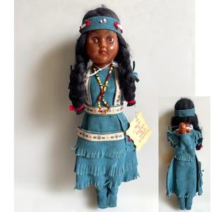 Può includere: Bambola nativa americana vintage con capelli neri, indossa un vestito in camoscio verde acqua con frange e una collana di perline. La bambola ha una fascia verde acqua con accenti rossi. Un'etichetta recita "Made in America by Native Americans."