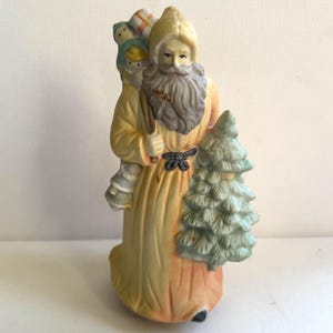 May include: A ceramic Santa Claus figurine in a long yellow robe, carrying a child on his back and a small Christmas tree. The figurine has a long gray beard and is holding a wooden staff. Presents are on the child's back. The figurine is painted with soft colors.