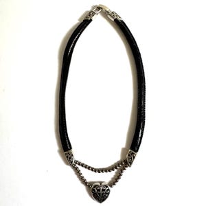 May include: A black leather necklace with a silver heart pendant. The necklace features a double strand design with a heart-shaped pendant at the center. The pendant has an ornate, openwork design. The necklace is approximately 16 inches long.