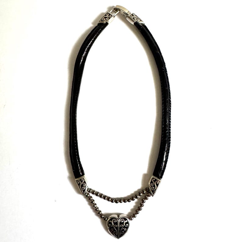 May include: A black leather necklace with a silver heart pendant. The necklace features a double strand design with a heart-shaped pendant at the center. The pendant has an ornate, openwork design. The necklace is approximately 16 inches long.