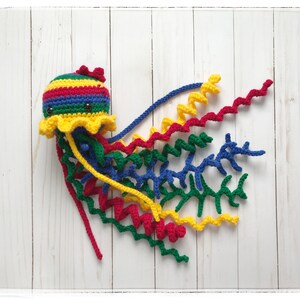 May include: A colorful, handmade crochet jellyfish toy. The jellyfish has a striped head in red, yellow, blue, and green, with a yellow frill and multiple curly tentacles in red, yellow, blue, and green. The toy has two small black eyes and a red bow.