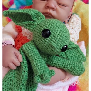 Puede incluir: Una muñeca bebé durmiente sostiene una criatura verde tejida con grandes ojos negros y largas orejas. El bebé está envuelto en una manta rosa, con una colorida manta tie-dye en el fondo. El texto "April Carrine Creates" es visible.