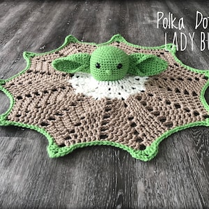 May include: A crocheted baby blanket in brown, white and green with a crocheted Yoda character in the centre. The blanket has a starburst shape with a scalloped edge. The text "Polka Dots & LADYBUGS" is visible in the top right corner of the image.