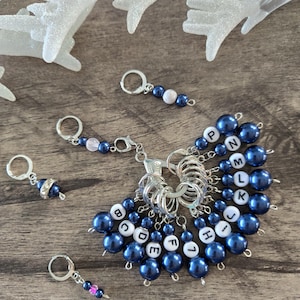May include: A collection of silver-toned metal stitch markers featuring dark blue pearl-like beads and white alphabet beads with black letters. The set includes individual markers with letters B through P, alongside several accent markers with pink or white beads.