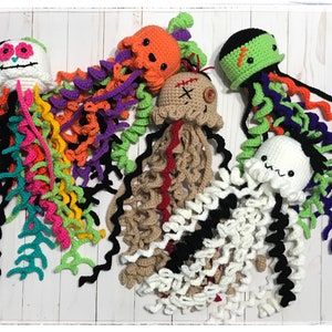Può includere: Una collezione di meduse all'uncinetto fatte a mano in vari colori. Ogni medusa presenta un design unico, tra cui un teschio di zucchero, una zucca arancione, un mostro di Frankenstein e una medusa bianca con i capelli neri. I tentacoli sono di colori vivaci.
