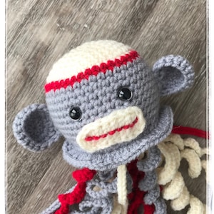 May include: A handmade crochet sock monkey with a gray body, cream-colored head, and red accents. The monkey has black button eyes, gray ears, and a cream-colored mouth with red stitching. It has a red and cream-colored tail.