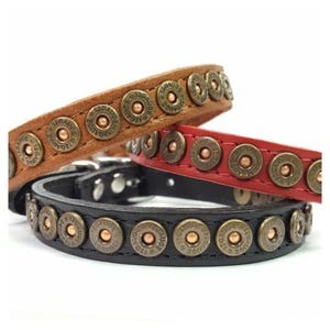 May include: Three leather collars in tan, red, and black, each featuring a row of brass-toned metal studs. The studs are circular, embossed with the text 12 GAUGE, and have a central copper-colored rivet. Each collar has a metal buckle.