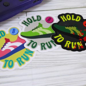 May include: Three colorful stickers with the text "HOLD TO RUN" and a sneaker graphic. The stickers are in yellow, pink, green, and black. A purple game console is in the background.