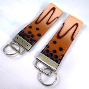 May include: Two keychains with a boba tea design. The keychains have a light brown background with dark brown boba pearls and a dark brown wavy design. Each keychain has a silver metal clasp and a silver key ring.