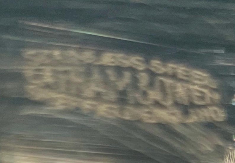May include: Blurred image of text that reads "STAINLESS STEEL GRAY RAVEN JEWELERS ENGLAND". The text is in a light color against a darker background.