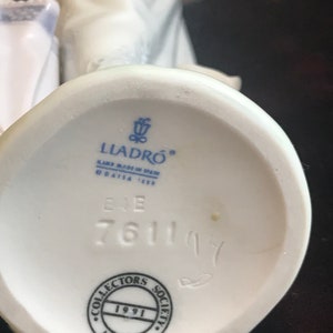 May include: White porcelain figurine with a blue and white dress and a white umbrella. The figurine is marked with the Lladro logo, "Hand Made in Spain", and the number "761114". The base of the figurine also has a black circle with the text "Collectors Society 1991 Lladro".