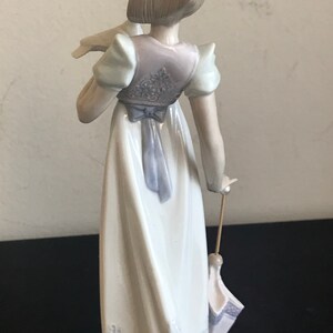 May include: A porcelain figurine of a girl in a white dress with a blue sash and a white lace trim. She is holding a white bird in her left hand and a closed umbrella in her right hand.