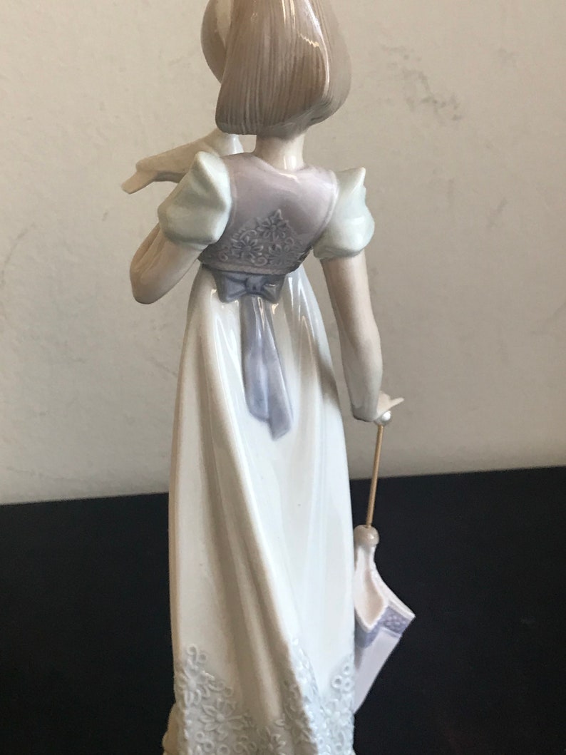 May include: A porcelain figurine of a girl in a white dress with a blue sash and a white lace trim. She is holding a white bird in her left hand and a closed umbrella in her right hand.