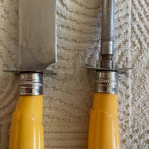 May include: Close-up of a carving knife and a sharpening steel. The knife has a silver blade and a yellow handle. The sharpening steel has a silver rod and a yellow handle. Both have silver accents.
