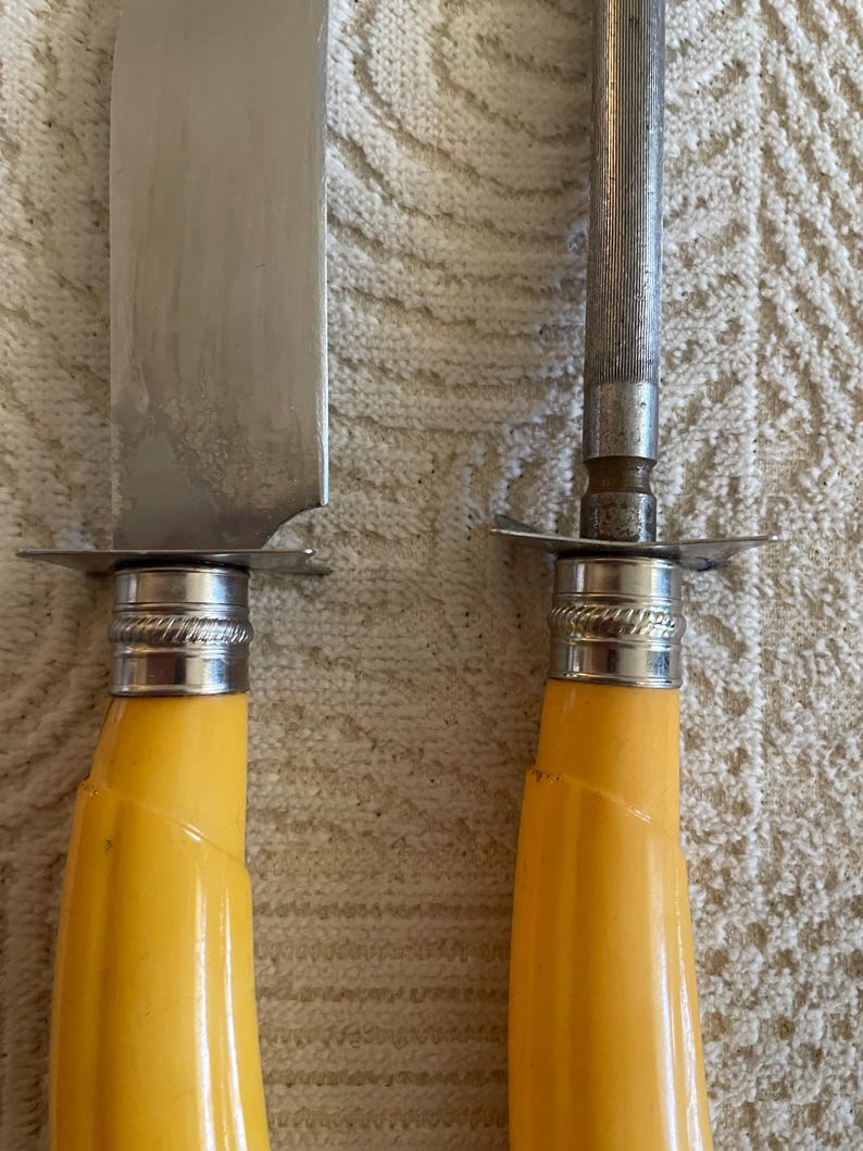 May include: Close-up of a carving knife and a sharpening steel. The knife has a silver blade and a yellow handle. The sharpening steel has a silver rod and a yellow handle. Both have silver accents.